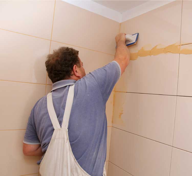 Professional bathroom renovation contractor applying grout to beige ceramic wall tiles during a detailed bathroom renovation project, showcasing expert craftsmanshi