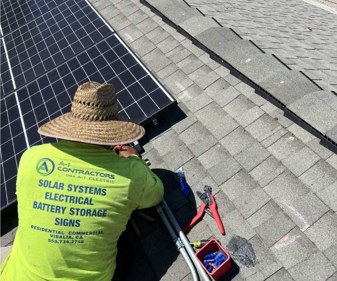 Solar technician wearing a safety hat and yellow shirt using wire cutters to install rooftop solar panels on asphalt shingles during a sunny day